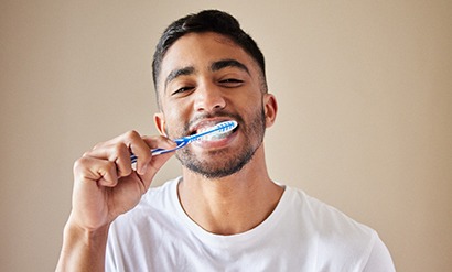 Man in white t-shirt brushing teeth in front of tan background