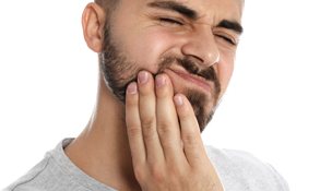 A young man with a beard holding his jaw and cringing in pain because he needs an emergency dentist